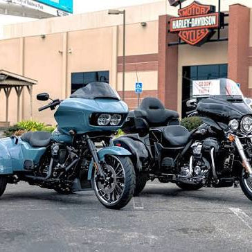 Two Harley-Davidson trikes parked in a dealership lot: a light blue trike with a sidecar on the left and a black trike on the right, with a Harley-Davidson building in the background.